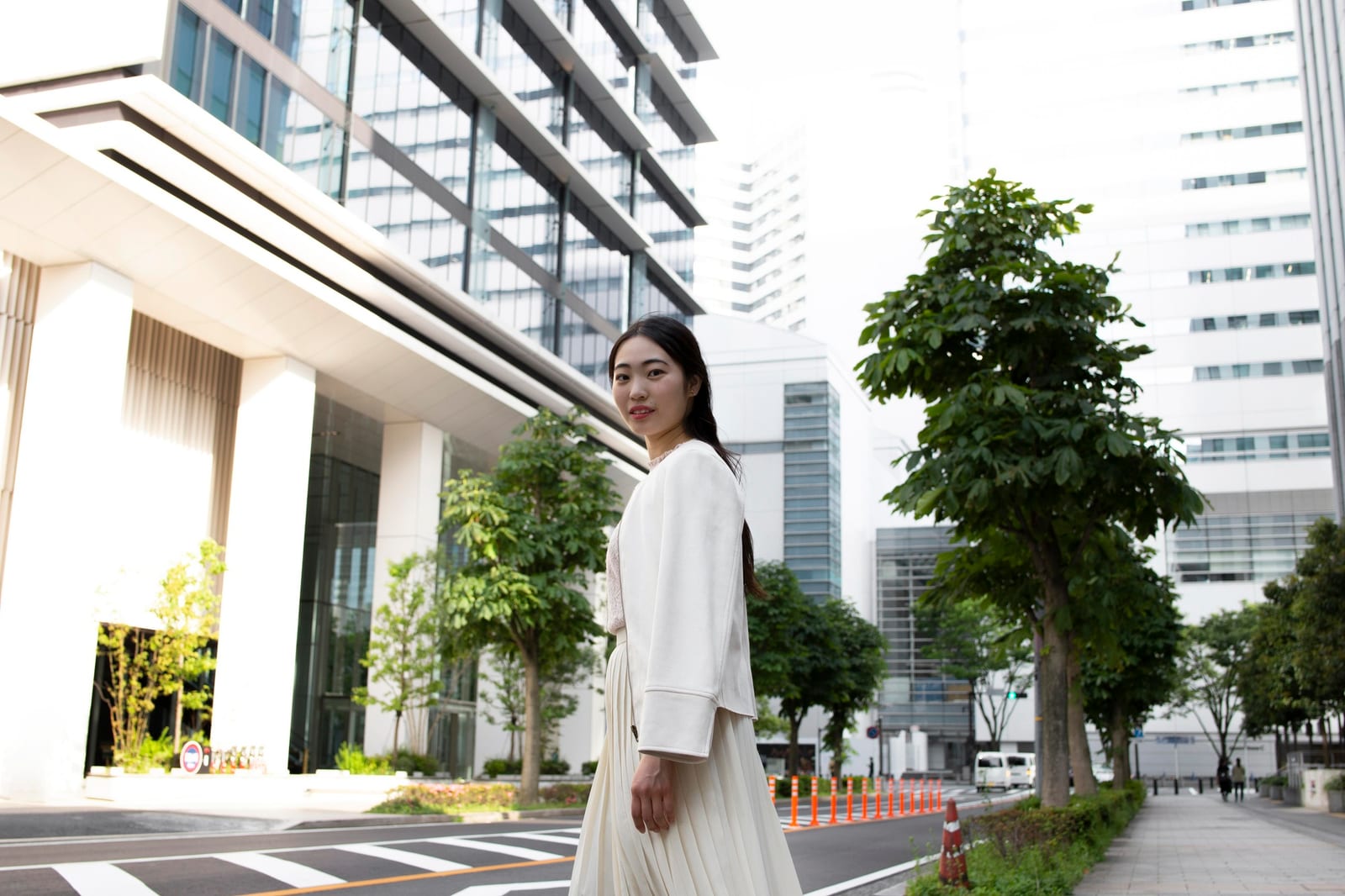 白い服装の若い女性が、近代的なビジネス街の横断歩道に立っている